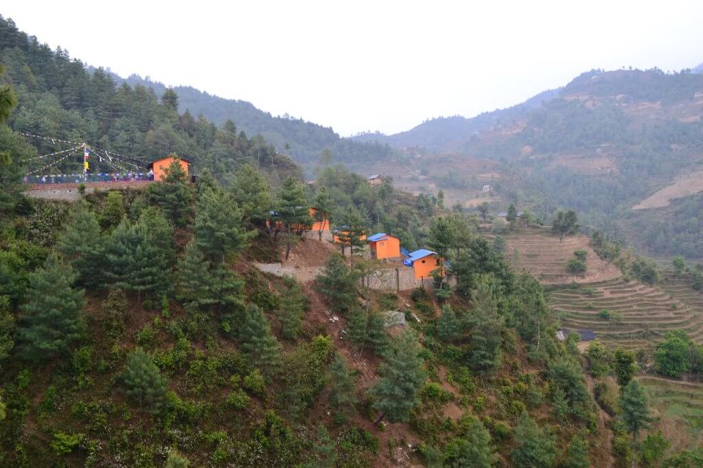The school buildings hug the mountain slope. Bright blue roofs, golden orange walls and colourful prayer flags over the playground show brightly through tall trees.