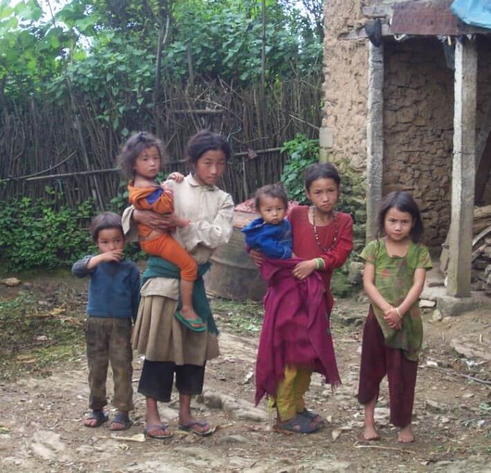 Six children between two and fourteen years old, one barefoot and ragged, are standing together on rough ground near a farm house. The older children are carrying the young ones.