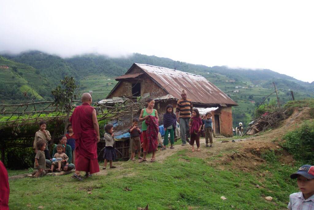 The family are poorly dressed. Their home is built from piled stone, wood and a metal roof. A distant view of hills and farm buildings is obscured by low clouds.