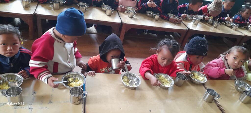 Children sit on cushions beside benches to eat their school breakfast of corn porridge. The children wear various warm outer clothes, hats, coats and jumpers because the morning is cold.