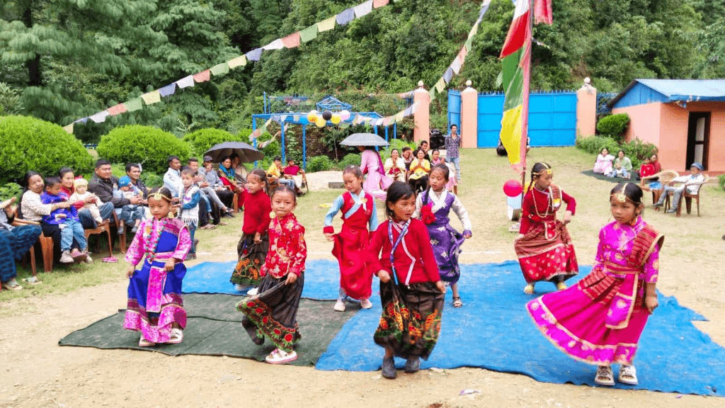 Eight girls in colourful traditional costumes are giving a dance performance in the playground for their parents who sit watching.