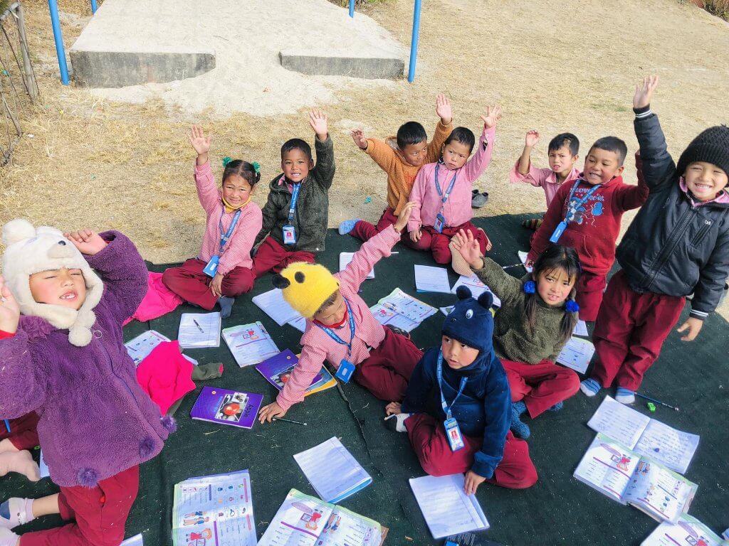 A class of young children some wearing hats and coats over their uniforms are seated on a mat in the playground working on colourful Nepali school textbooks.