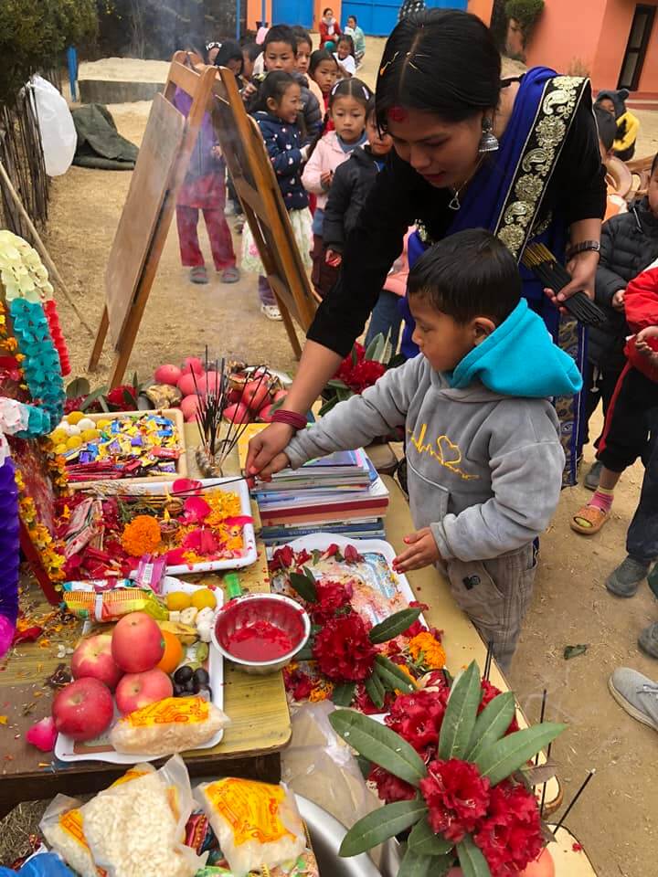 A boy stands beside a display of fruits and flowers, his teacher helps him to light incense as an offering. The rest of the class watch and wait for their turn.