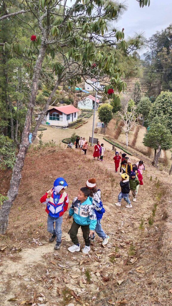 A stream of children and adults climb a stony hillside, behind them is a view of different species of trees, laid out planting beds and small attractive buildings.