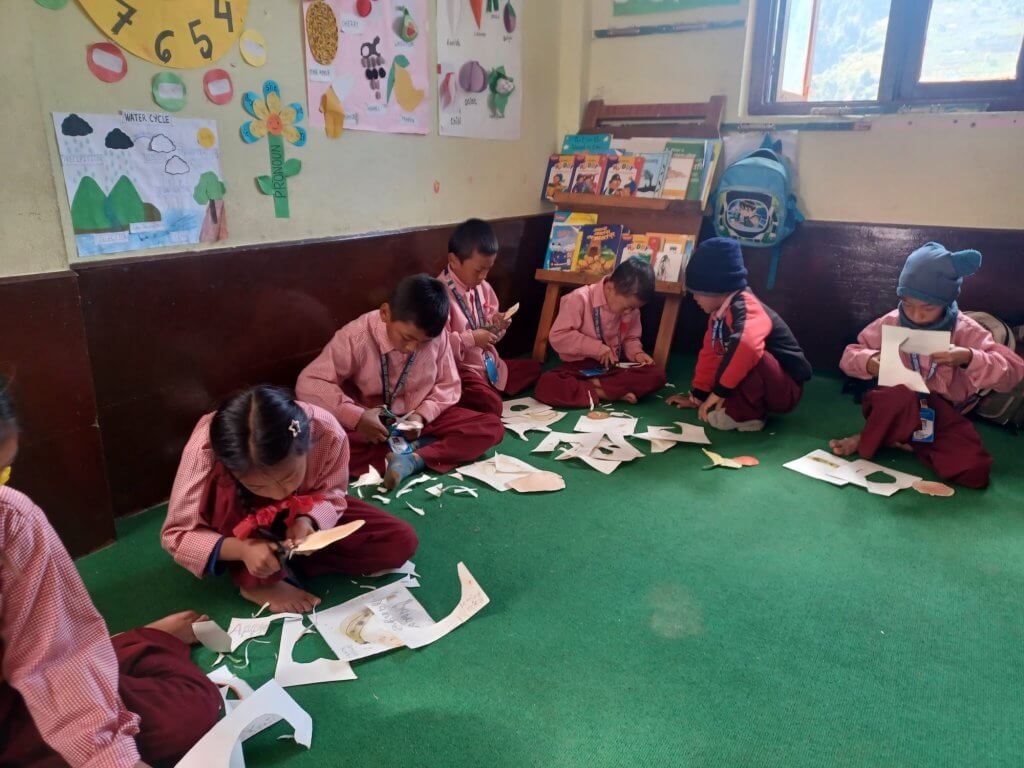 Class one are sitting on their classroom floor cutting out drawings of fruit that they have made for an art project. The children are in neat uniforms but all have bare feet, because the room is cold two children have hats on.
