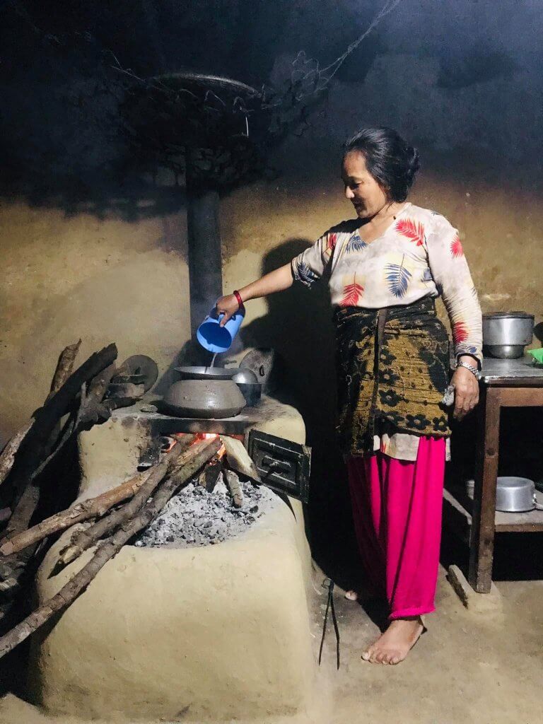 A women stands pouring water from a plastic jug into a large cooking pot atop her stove. The stove is raised and encased by a smoothly finished unfired clay base, with a bowl shaped depression to catch spilling ash. An attached chimney takes most of the smoke from the room.