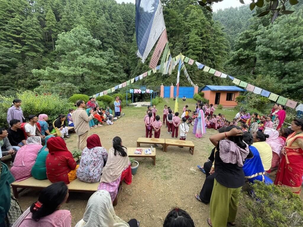 The edge of the playground is lined with families watching and listening to their children perform. Huge trees frame the view of a sturdy blue gate to the school grounds.