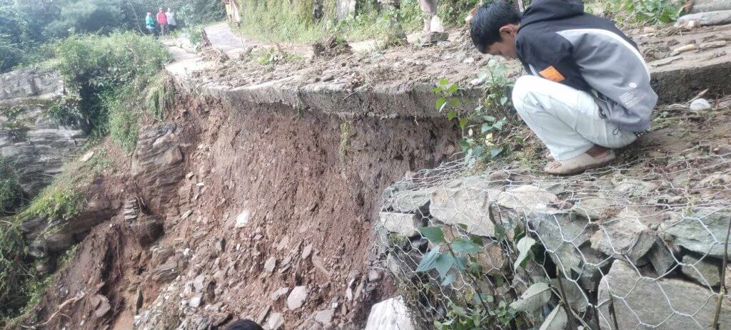 A boy crouches on the edge of a badly damaged road and looks down the steep drop.