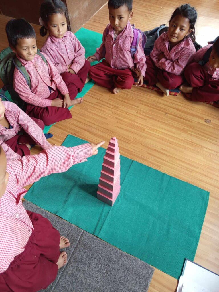 Seven children are sitting on the floor, one child points to the top of a tower she has built, the other children are looking with interest and concentration.