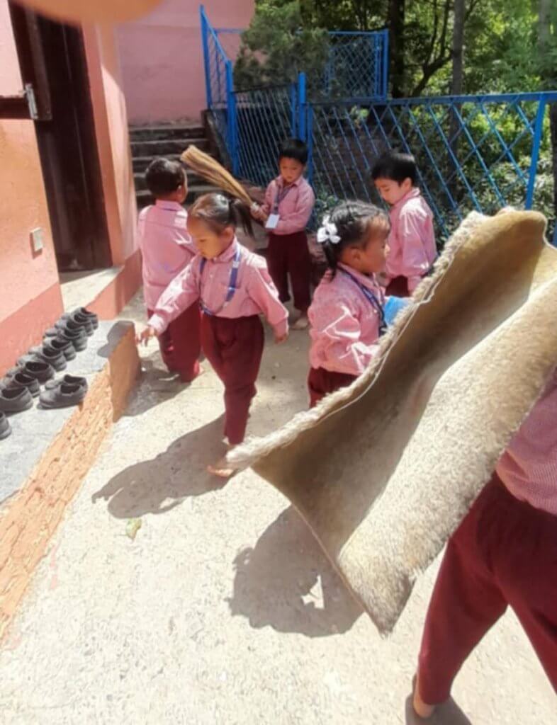 A group of children are busy on the path outside their classroom, they have arranged the shoes neatly beside the wall and are sweeping the path. One child is shaking the doormat.
