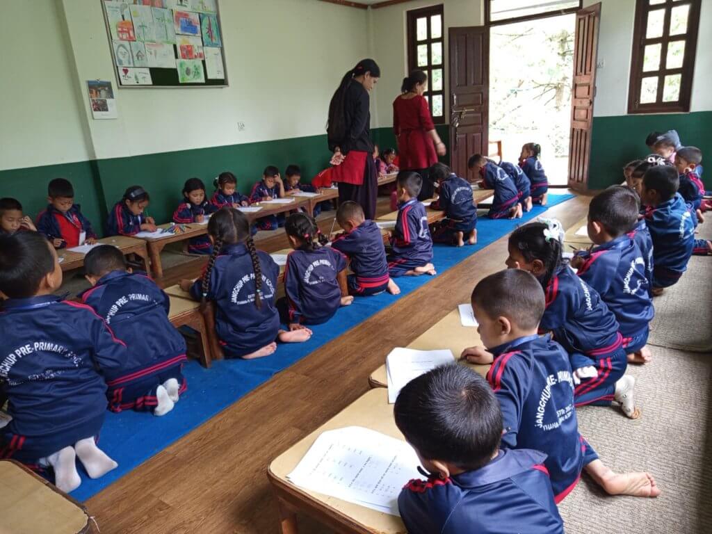 Three rows of children in blue track suit style uniform kneel at benches in a classroom. They are taking a written test on a sheet of paper. The school door is open to the fresh air there are trees outside.