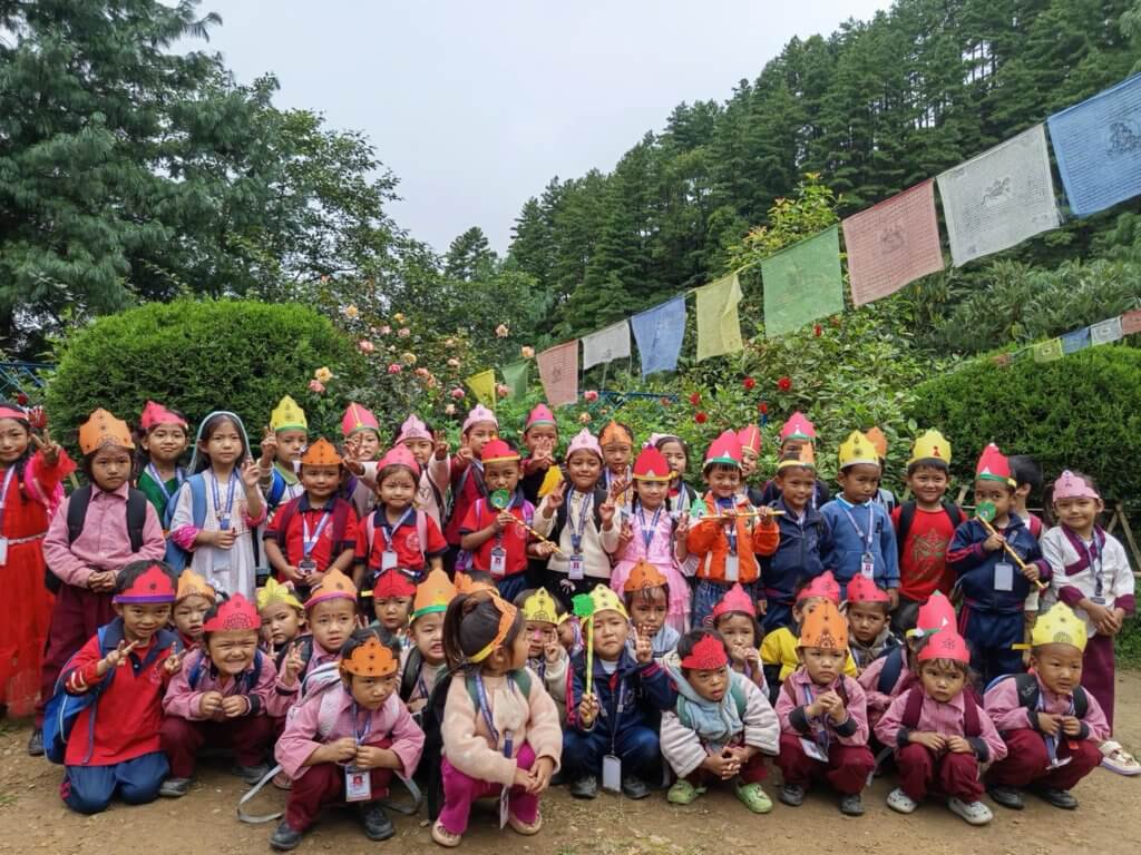 A group image of all the children dressed in colourful headdresses with a background of trees and Buddhist prayer flags.