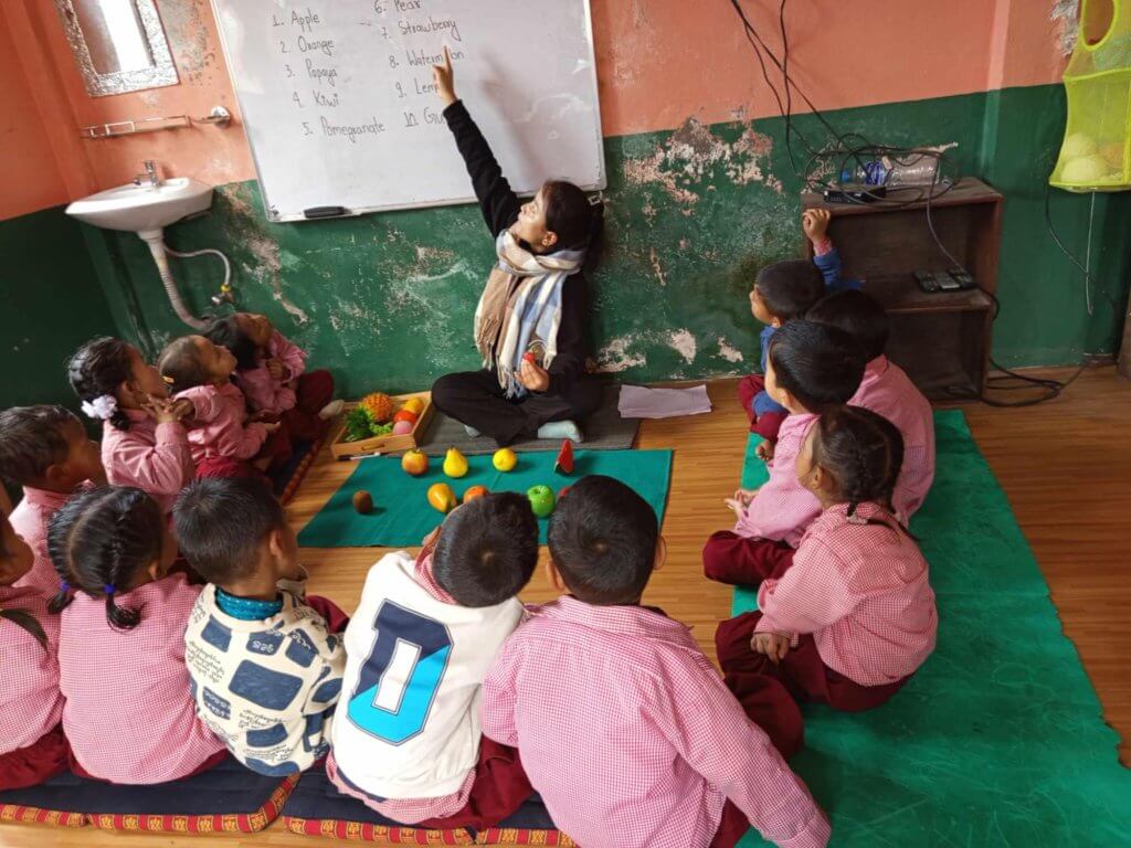 Whilst sitting on the floor, teacher stretches up and points to the writing on the board. There are ten fruit models on the floor. The children are looking at the English names of the fruit on the board.