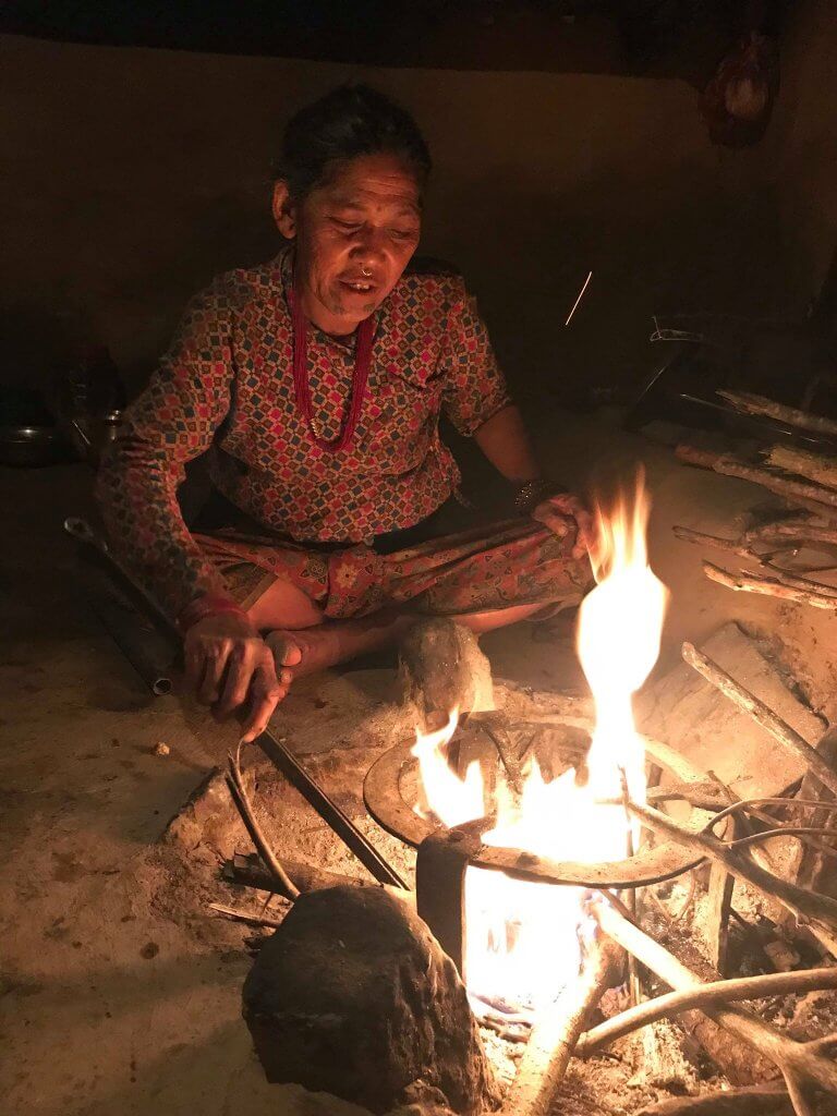 A woman is seated cross-legged on the earth floor tending an open fire pit which has a simple metal trivet for supporting a cooking pot.