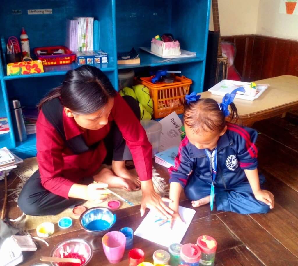 A female teacher is sitting on the class room floor with different coloured paints arranged in front of her. Teacher is helping a young girl with a painted palm to press her hand to a piece of paper.