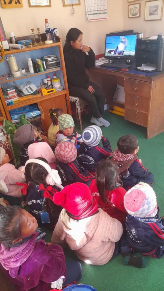 Young children wearing hats and coats sit on the office floor, looking up at the laptop screen on the desk. They are watching cartoons; their teacher sits beside the laptop.