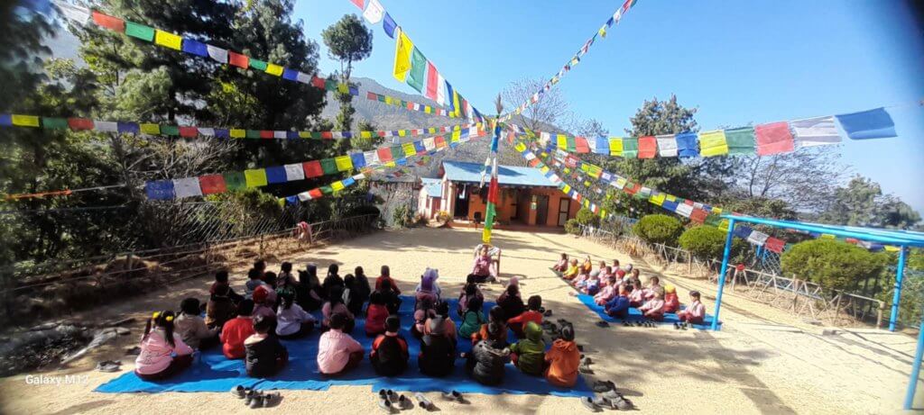 About fifty children are sitting grouped on mats in the playground. The roof of the school is blue like the sky, its walls are golden orange. Above them flutter strings of colourful Buddhist prayer flags.