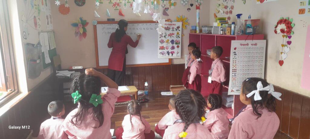 Teacher is writing on the board children are standing or sitting watching .A child has her hand ready to answer a question. The room is decorated with colourful crafts and posters.