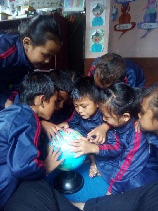 A group of six children are clustered around a globe, their hands are touching the globe with fingers spread. They are looking at it intently with pleasure and fascination.